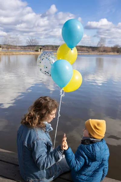 Молодая Женщина Ребенок Сидят Берегу Озера Держа Желтые Синие Воздушные — стоковое фото