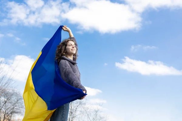 stock image an adult girl of 18 years old, holds a Ukrainian yellow-blue flag behind her back against the sky, looks ahead, in the hope of a brighter future. Ukrainians are against the war. Support Ukraine