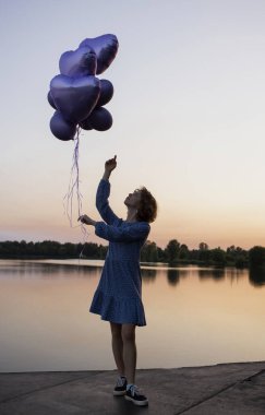 Elbiseli genç, narin bir kadın elinde bir sürü balon tutuyor, rüyalarında gün batımında gölün kenarında duruyor. fantezi, özgürlük, neşe, tatiller, pozitif. Kızın doğum günü.