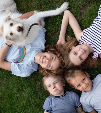 Farklı yaşlardaki çocuklar, erkek kardeşler, çimenlere uzanıp kameraya bakıyorlar. Dost canlısı, mutlu kızlar ve oğlanlar ve favori beyaz köpekleri. Yaz tatili, tatil, birlikte eğlenmek.