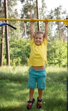 boy of 5-6 years old hangs on his hands on the horizontal bar in the park. Active lifestyle, fun activities for children in the summer outdoors, sports for physical development clipart