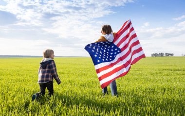 Kadın ve çocuk, anne ve oğul, neşeyle Amerikan bayrağını taşıyan yeşil çayır boyunca ilerliyorlar. Vatansever bayram, Bağımsızlık Günü. Amerikalı olmaktan gurur duyuyorum. Özgürlük, ulusal kimlik