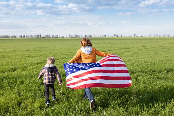 一个女人和一个孩子欢快地跑过一片绿色的草地 美国国旗在他们身后飘扬 作为一个美国人感到自豪 国家自由象征 美利坚合众国独立日 — 图库照片