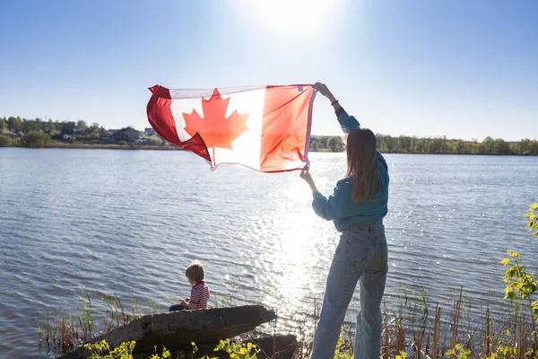 in hands of woman, Canadian flag is fluttering on a sunny day against the sky and a lake, a child is sitting nearby. Travel, holidays, immigration. Canada Day. Pride, freedom, national symbol