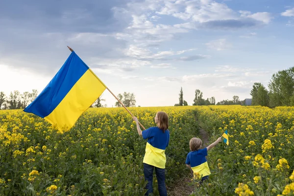 菜種畑の間で手にウクライナの旗を持つ黄青のTシャツの母親と息子 愛国心の教育です ウクライナの独立記念日 平和のための子供たち 戦争をやめろ ウクライナ人であることへの誇り — ストック写真