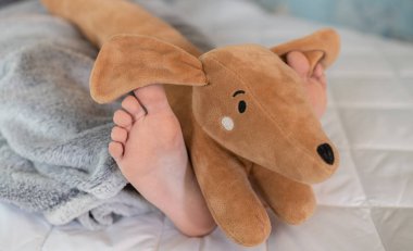 Çocuğun çıplak ayağı yatağın üzerinde ve yumuşak tüylü oyuncak bir köpek. İyi sabahlar. Rahat bir uyku yeri, en sevdiğin oyuncağınla zevkle uyu.