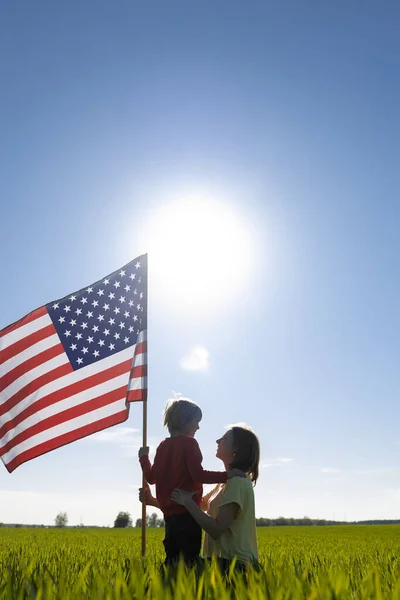 轮廓中的妇女和儿童 母亲的儿子在绿地里互相望着 在灿烂的阳光下举着美国国旗 爱国假日 独立日 作为一个美国人感到自豪 民族特性 — 图库照片