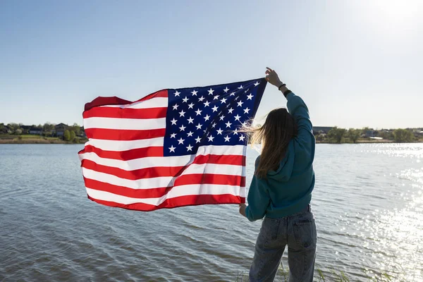 在阳光明媚的日子 在天空和河流的衬托下 举着美国国旗的女人 自由的国家象征 美利坚合众国的独立 — 图库照片