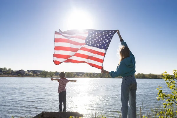 女人手里拿着美国国旗 美丽的阳光照耀着她 孩子高兴地把胳膊伸向一边 爱国假日 独立日 作为一个美国人感到自豪 国家自由的象征 旅行假期 — 图库照片