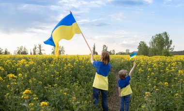 Kadın ve bir çocuk, kolza tohumu tarlasında ellerinde Ukrayna bayrakları olan sarı ve mavi tişörtler giyiyorlar. Vatanseverlik eğitimi. Ukrayna 'da Bağımsızlık Günü. Ukraynalı olmaktan gurur duyuyorum.
