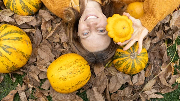 快乐的微笑着的女人躺在大大小小南瓜中间的一堆堆干树叶上 其中的一片叶子手牵着脸 选择焦点 顶部视图 美丽多彩的黄色 橙色秋天的气氛 — 图库照片
