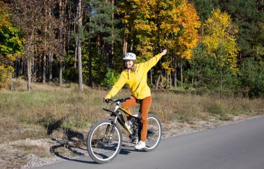 Genç bir kadın güneşli bir günde sonbahar parkında bisiklet sürüyor. Özgürlük ve neşe hissi. Aktif yaşam tarzı. Doğanın portakal rengi sonbahar arka planında eğleniyor.