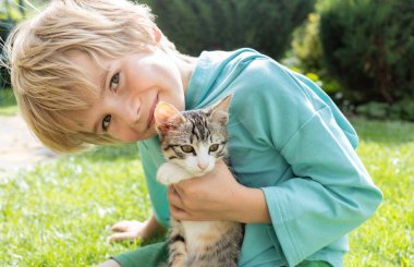 Yavru kedi, güneşli bir günde 7 yaşında tatlı bir çocuğun kollarında oturur. Kedi günü. Evcil bir hayvanın yanında mutlu bir çocukluk. pozitif bir hayvan ve çocuk arkadaşlığı ortamı