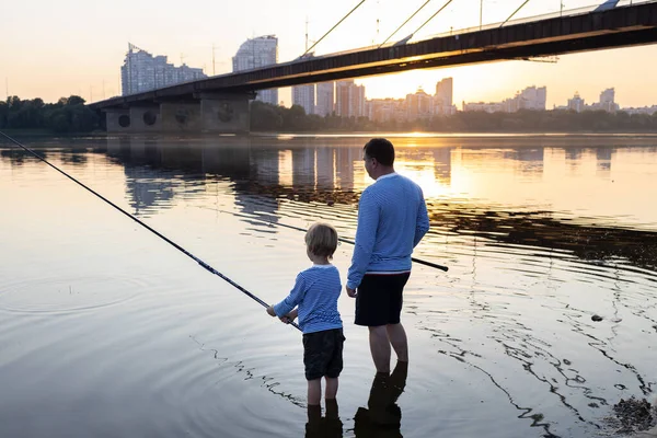 Father and son are fishing together standing with fishing rods in the water of the river at sunset. father\'s day concept. Family fun pastime for the weekend. Hobby for child and parent