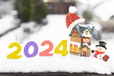 Mutlu yıllar 2024. Kardaki numaralar, kardan adam figürleri, rahat bir kulübe, Noel Baba şapkası. Tebrik kartı kış tatili havasında. Noel, yeni yıl ve kış tatillerini kutlamak için konsept