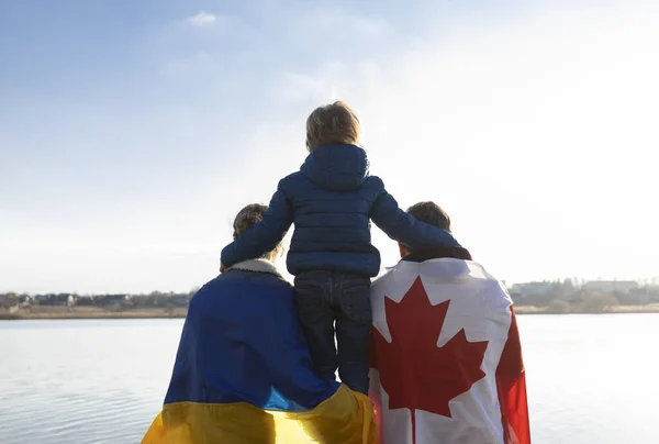 woman with Ukrainian flag, man with Canadian flag behind back and child , sitting with backs together in nature . International support and assistance to Ukraine. Problems of refugees and immigrants