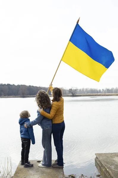 boy and two women, mother with children, next to lake with Ukrainian flag. Family, unity, refugees, support. stand with Ukraine. stop the war. need for international support and assistance to Ukraine