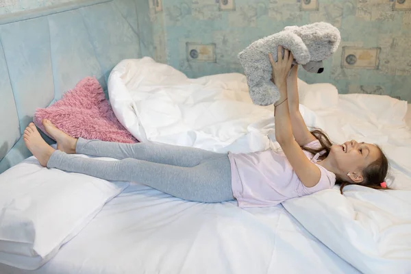 stock image 8-year-old girl lies in bed after waking up, lifting a teddy bear up in her hands, cheerfully placing her bare feet on the pillow. cozy rest in the bedroom, children's games with soft toys