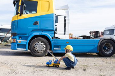 boy in construction helmet with toy car plays with interest while sitting on construction site in front of dump truck cabin. Game in the profession. Builder's Day. Interesting games for boys clipart
