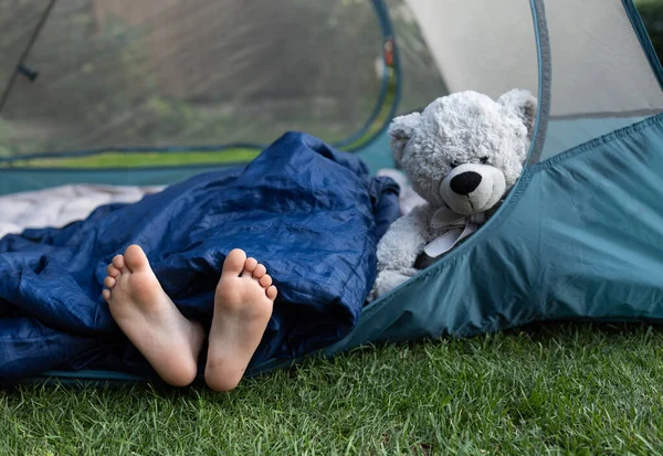 cute bare feet of a child sticking out of a sleeping bag. A child with his favorite toy, a teddy bear, sleeps in a tent. Healthy lifestyle, little tourist, family activities, hiking in summer