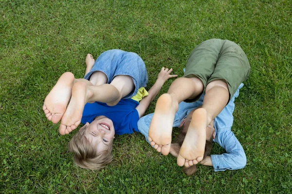 2人の陽気な裸足の子供が足で草の上に横たわっている 公園の兄弟や友人たちを苦しめる 夏の気分 ハッピーケアフリーの子供時代 パンフレット ロイヤリティフリーのストック写真