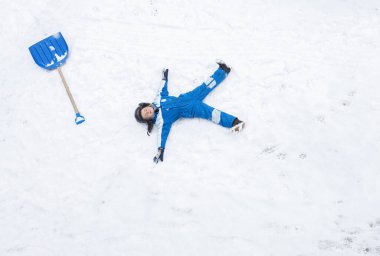 Mavi kışlık takım elbiseli çocuk kar fırtınasından sonra kar temizledi ve yakınlarda büyük bir kürekle dinlenmek için karın üzerine uzandı. Kış zamanı, kötü hava, soğuk, karla oynamak, şımartma, mizah. Yukarıdan görüntüle