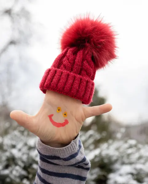 smiling face is drawn on the child palm, and a warm red knitted hat is put on his fingers. It\'s time to warm up and enjoy winter walks. happy childhood, winter holidays, pampering
