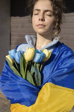 Ukrayna bayrağına sarılmış düşünceli genç bir kadın elinde bir buket sarı ve mavi lale tutuyor. Bağımsızlık Günü. Ukrayna için dua et. Ukrayna 'yı Destekle.