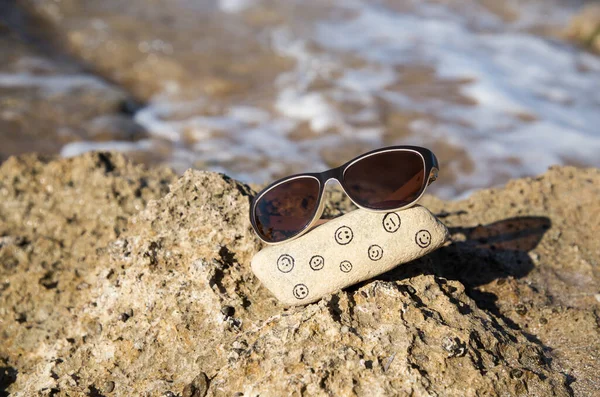 natural stone with funny and sad faces painted on it and sunglasses on the sea coast. Hello summer, sun, vacation, positive emotions, travel. Choose your mood, be positive, digital detox