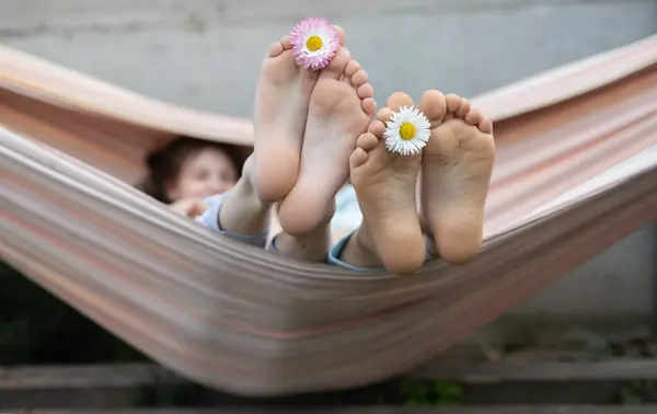two barefoot children relax together sitting in a hammock. Lifestyle. Between the toes of their bare feet they inserted daisy flowers. Have fun, enjoy life. Summer holidays
