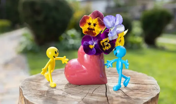 黄色と青のミニチュアのおもちゃの人間の数字は 晴れた日のパンジーの花束と心臓型の花瓶の近くに 恋愛のカップルとロマンス 愛と優しい感情の概念 ロイヤリティフリーのストック写真
