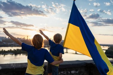 Ukrayna bayrağı taşıyan anne ve oğlu, Kyiv 'deki Dinyeper Nehri ve gökyüzünün arka planına karşı çatıda ayakta duruyor. Ukrayna 'daki savaşa dikkat çeken vatanseverlik. Bağımsızlık Günü.