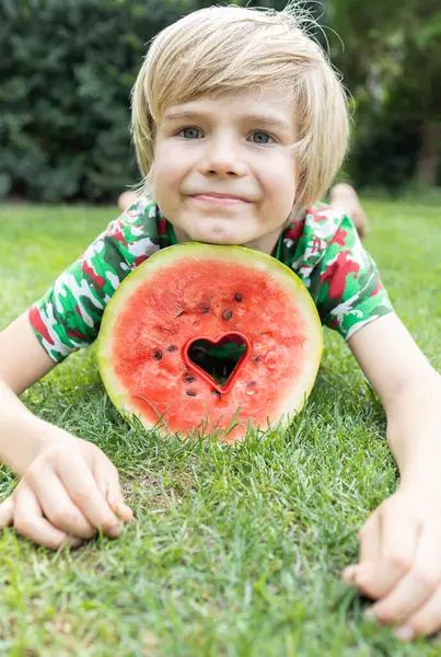 round piece of watermelon with a carved heart inside and the face of a boy lying on the grass. Summer mood. Proper nutrition. Vegetarian food. Favorite juicy vitamin food