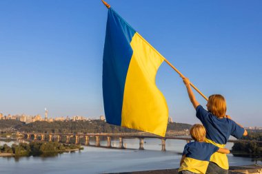 Sarı-mavi bayraklı kadın ve çocuk Kyiv 'deki Dinyeper Nehri' ne karşı bir evin çatısında dikiliyorlar. Vatanseverlik, Ukrayna 'daki savaşa dikkat çekmek. Ukrayna 'yı destekleyin. Bağımsızlık Günü