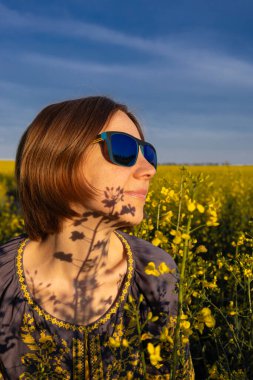 Sarı kolza tohumu tarlasının arka planına karşı şehvetli kadın portresi ve mavi gökyüzü, çiçeklerin yüzünde gölgeler. Mavi güneş gözlüklü ve Ukrayna işlemeli bluzlu bir kadın.