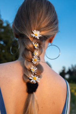 Sarışın kadın sırtı dönük ve saçları papatya çiçekleriyle örülmüş bir şekilde ayakta duruyor. Doğanın tadını çıkar, yaz havası.