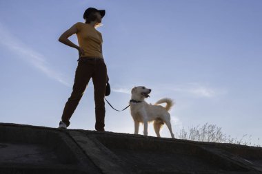 Gün batımında köpeğiyle gökyüzüne bakan bir kadının tam boy silueti. Sevdiğin hayvanınla yürü. Dört bacaklı bir arkadaşla iletişim kurmanın keyfi.