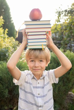 Sevimli meraklı çocuk kafasının üstünde bir yığın kitap ve bir elma tutuyor. Okula dönüş, ilginç bir eğitim, kitap severler günü. Dijital detoks. Kitap Günü. Kitapların değeri