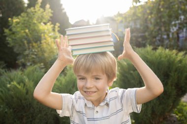 Sevimli meraklı çocuk bir yığın kitabı kafasında tutuyor ve dengelerini tutturuyor. Okula dönüş, ilginç bir eğitim, kitap severler günü. Dijital detoks. Kitap Günü. Kitapların değeri. Arkaplan ışığı, yumuşak odak