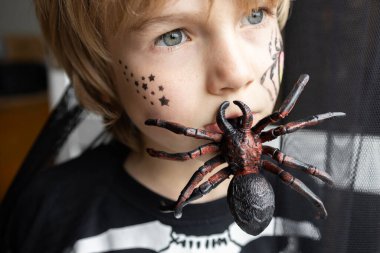 Çocuğun yüzüne yakın çekim, dişinde büyük bir örümcek tutuyor. Çocuk başka yere bakıyor. Cadılar Bayramı. Şaka mı şeker mi?