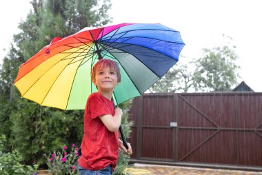Gökkuşağı şemsiyeli çocuk yaz duşunun altında duruyor. Bulutlu bir günde, 7 yaşında bir çocuk çok renkli bir şemsiyenin altında duruyor ve etrafına yağmur yağıyor. Mevsimlik sevinçler, bulutlu yağmurlu hava.
