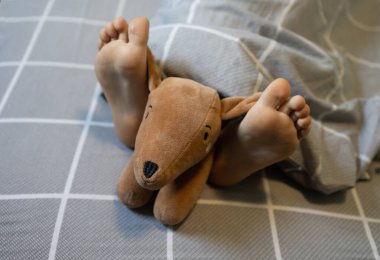 Çocuğun çıplak ayaklarıyla battaniyenin altındaki yatağın üzerinde yumuşak ve komik bir oyuncak köpek yatıyor. İyi sabahlar. İyi geceler. Rahat bir uyku yeri, en sevdiğin oyuncağınla zevkle uyu.