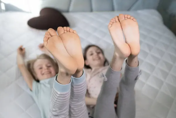 stock image Two cheerful children playing around while lying on bed, cheerfully raised up bare feet. focus on children's toes and heels. friends or siblings at home having fun on parents' big bed, family joy