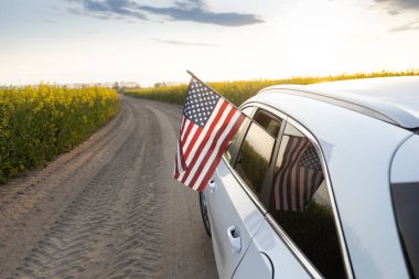 Amerikan bayrağı, batan güneşe ve açan kolza tohumu tarlasına karşı off-road yapan araba camından sarkıyor. ABD Bağımsızlık Günü 4 Temmuz. Gurur, özgürlük, vatanseverlik, demokrasi. Ülkeyi dolaş.