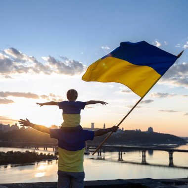 Ukrayna 'nın büyük bayrağıyla babamın omuzlarında oturan çocuk Dinyeper seti üzerindeki bir evin çatısında ve Kyiv' in manzarasında dikiliyor. Vatansever eğitim. Savaşı durdurun. Bağımsızlık Günü