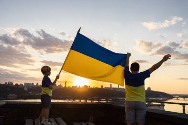 Sarı ve mavi tişörtlü çocuk ve adam büyük bir Ukrayna bayrağı taşıyan Dinyeper nehrinin önündeki bir evin çatısında ve Kyiv manzarasında dikiliyorlar. Vatansever eğitim. Bağımsızlık Günü