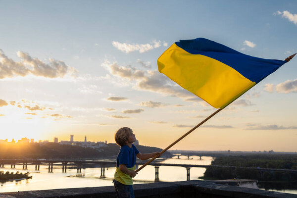 Мальчик машет большим украинским флагом против закатного неба и пейзажа города Киева. Украинский патриотический ребенок мечтает о мире для своей родины. Национальный символ свободы и независимости