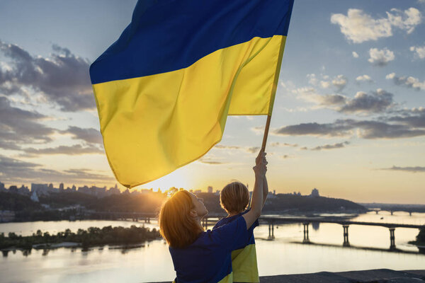 Мама и сын с большим украинским флагом стоят высоко на крыше дома напротив Днепра в Киеве и заката. Патриотизм, привлечение внимания к войне в Украине. День Независимости