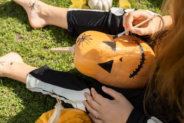 stock image child paints a scary grimace on a Halloween pumpkin with black paint. creative process, preparation for Halloween. Children's joys and entertainment.