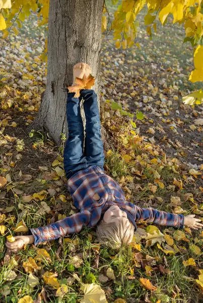 Çocuk yere yatar, ayakkabılarını çıkarır ve çıplak ayaklarını ağaç gövdesine kaldırır. pozitif atmosfer, mutlu çocukluk, doğadaki çocuklar, Hint yazı. Merhaba, Autumn. mevsimsel atmosferik fotoğraf